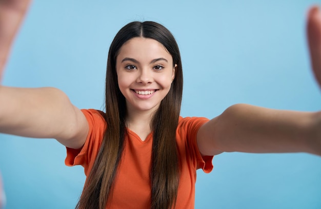 Ritratto del primo piano di una bella ragazza adolescente caucasica che tiene il telefono cellulare in mani tese sorridendo con un allegro sorriso a trentadue denti guardando la fotocamera isolata su sfondo di colore blu fotocamera pov