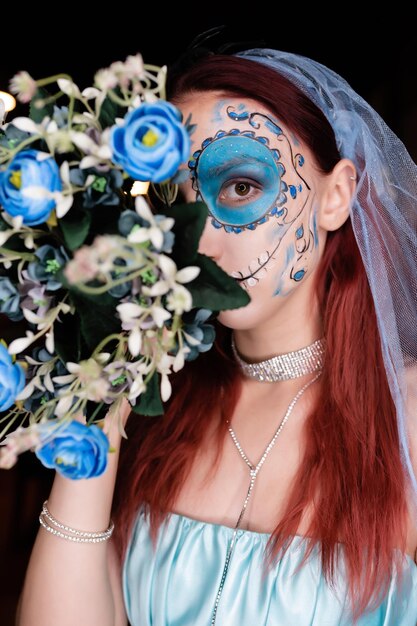 Closeup Portrait of a beautiful girl in a blue dress with artistic makeup on her face holding a bouquet of flowers Halloween mask Halloween concept nightclub masquerade