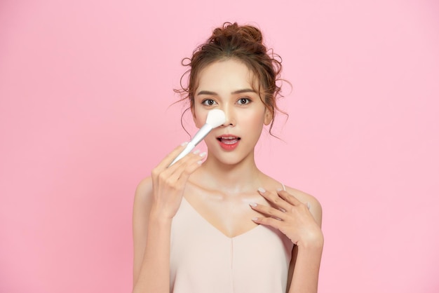 Closeup portrait of beautiful happy young girl with perfect skin is holding makeup brush in hand Isolated on pink