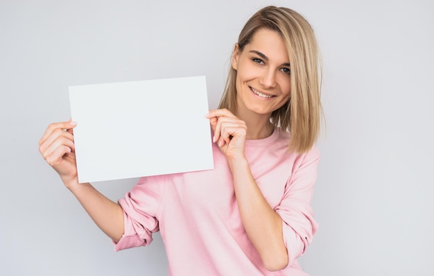 Closeup portrait of beautiful smiling blonde female wearing pink sweater posing with white blank paper with copy space for your advertisement information and looking at camera against white wall