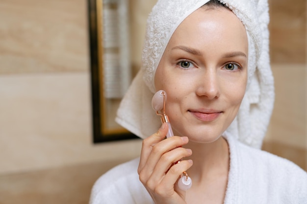 Closeup portrait of a beautiful woman massaging her face with a\
quartz gouache roller on the background of a mirror in the bathroom\
concept of cosmetic spa treatments and self care