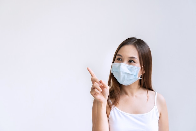 Closeup portrait of beautiful young Asian woman with protective face mask pointing with index finger upwards, isolated on white wall. Positive thinking concept. Copy space