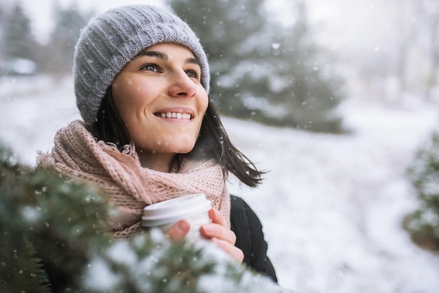 Closeup portrait of beautiful young smiling caucasian woman in winter jacket knitted beanie hat and scarf holding a cup of coffee or tea outdoors in park or forest on snowing day Winter mood