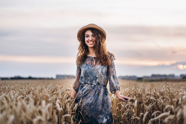 곱슬 머리를 가진 아름 다운 젊은 여자의 근접 촬영 초상화. 밀밭에 드레스와 모자 서있는 여자