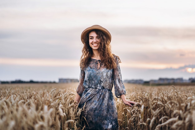 Closeup portrait of a beautiful young woman with curly hair. Woman in dress and hat standing in wheat field