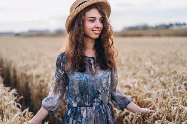 Closeup portrait of a beautiful young woman with curly hair. Woman in dress and hat standing in wheat field