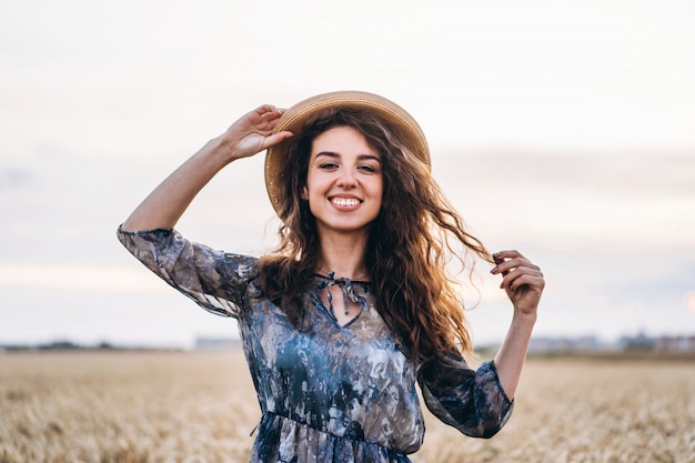 Closeup portrait of a beautiful young woman with curly hair. Woman in dress and hat standing in wheat field