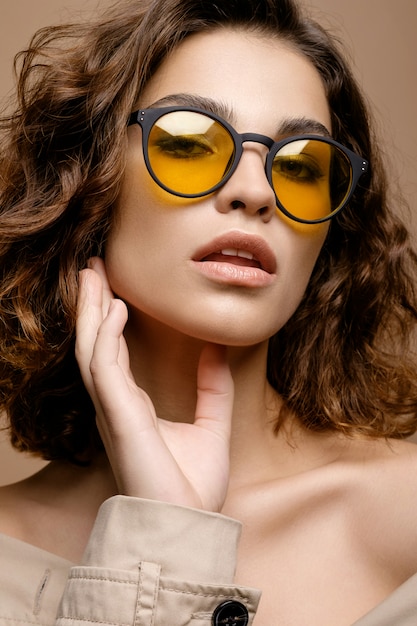 Closeup portrait of beauty fashion model with clean skin and curly hair in beige cloak stretch on beige wall, model in fashion glasses, bare shoulders