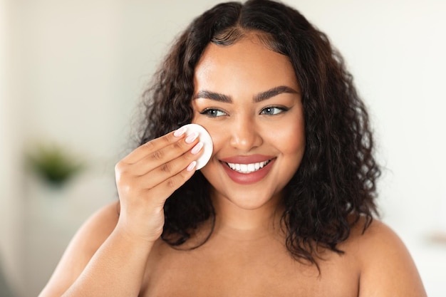 Closeup portrait of black plus size woman using cotton pad erasing make up from her face applying facial toner