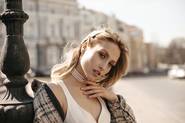 Foto il ritratto del primo piano della donna bionda splendida nel trucco bello che esamina la macchina fotografica all'esterno la signora attraente in vestito bianco e la giacca a scacchi posa alla piazza della città