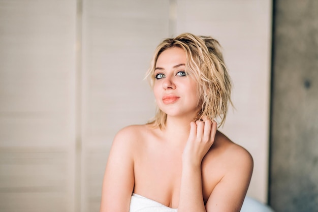 Closeup portrait of blonde naked woman with short wavy hair, full lips, beautiful eyes, perfect clean skin wrapped in white bath towel, being satisfied going to have spa massage in beauty salon.