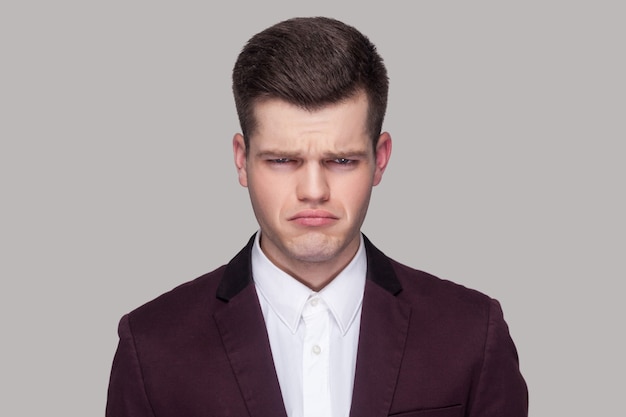 Closeup portrait of bored sad handsome young man in violet suit and white shirt, standing and looking at camera with bad mood. indoor studio shot, isolated on grey background.