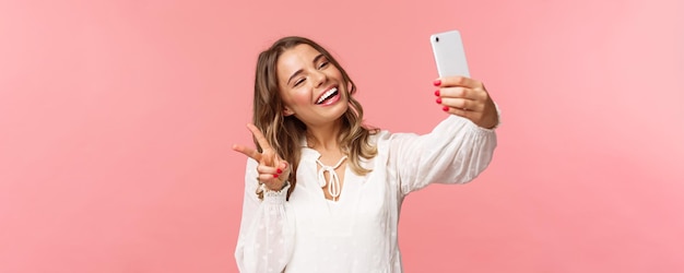 Il ritratto del primo piano della ragazza bionda femminile adorabile allegra in vestito bianco che prende il selfie sul telefono cellulare fa il segno di pace di kawaii mentre scatta la foto che cattura il fondo rosa del momento della primavera