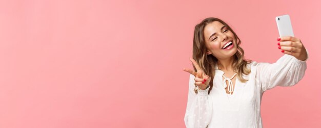 Closeup portrait of cheerful lovely feminine blond girl in white dress taking selfie on mobile phone make kawaii peace sign while take photo capturing spring moment pink background