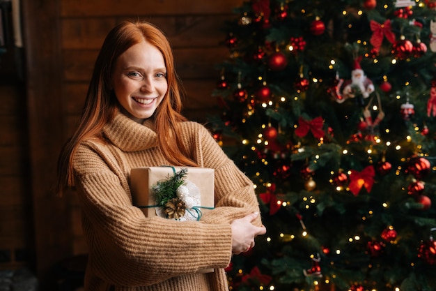 クリスマス ツリーの背景にクリスマス ギフト ボックスを保持している陽気な若い女性のポートレート、クローズ アップ