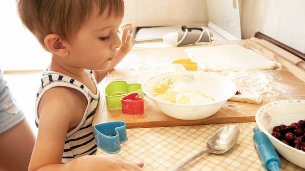 Closeup portrait of cute 3 years toddler boy standing on kitchen and cooking dough. Child baking and making breakfast