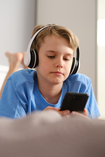 Closeup portrait of a cute boy in headphones A teenager with a smartphone lies on a bed at home