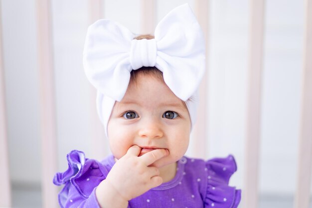 Closeup portrait of a cute and joyful baby girl in a crib in lilac clothes sucking her finger funny little baby at home baby products concept