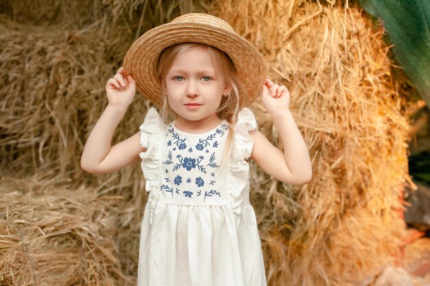 Closeup portrait of cute little blonde girl wearing light dress with embroidery and straw hat standing on hayloft on blurred background of haystack on sunny summer day Rural vacation concept