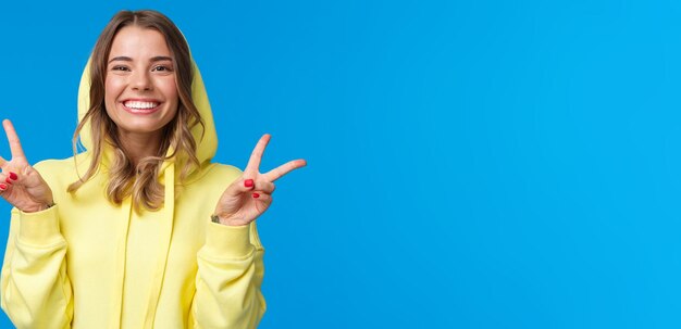 Closeup portrait cute silly blond european female in yellow hoodie showing peace gesture and smiling