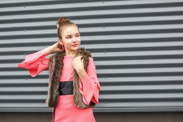 Closeup portrait of cute smiling little girl in pink dress and fur sleeveless jacket posing near gray corrugated striped wall