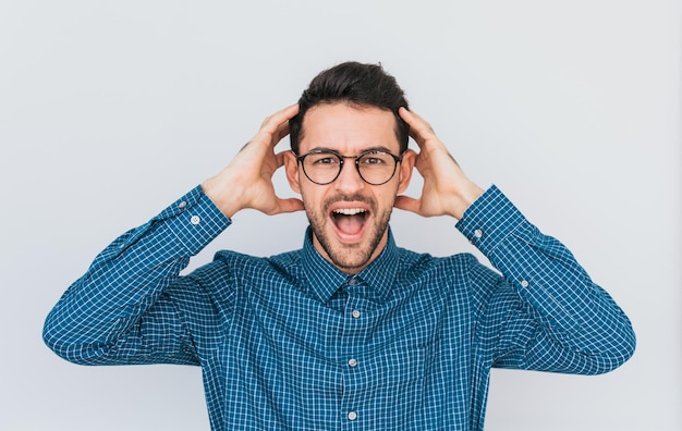 Closeup portrait of desperate stressed young handsome male wears glasses and blue shirt looking at camera in terror and shock keeping mouth wide open holding headOh no People and emotions concept