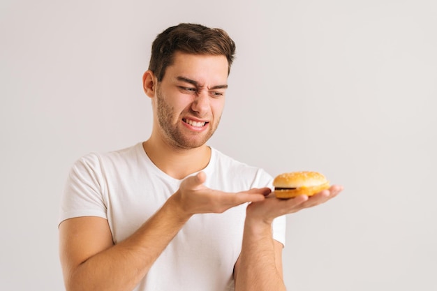 Ritratto del primo piano del giovane dispiaciuto con disgusto che indica l'hamburger difettoso su fondo bianco isolato