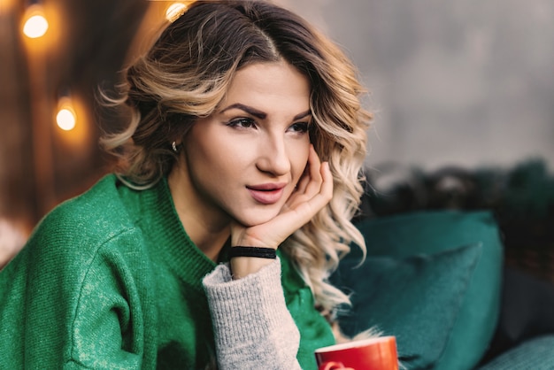 Closeup portrait of a dreaming blonde woman in a green sweater with a red cup in  hands. 