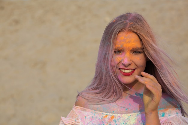 Closeup portrait of emotional young woman playing with exploding dry Holi paint at the desert. Space for text