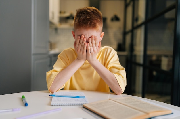 Photo closeup portrait of exhausted pupil boy tired from studying closed face with hands sitting at desk with paper notebook frustrated bored child schoolboy