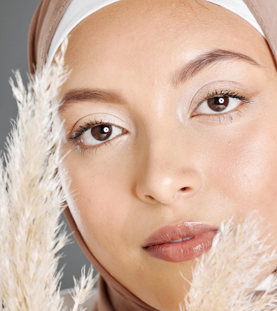 Closeup portrait face of beautiful young muslim woman wearing natural makeup and headscarf posing with pampas wheat plant against studio background Glowing modest arab covered in traditional hijab