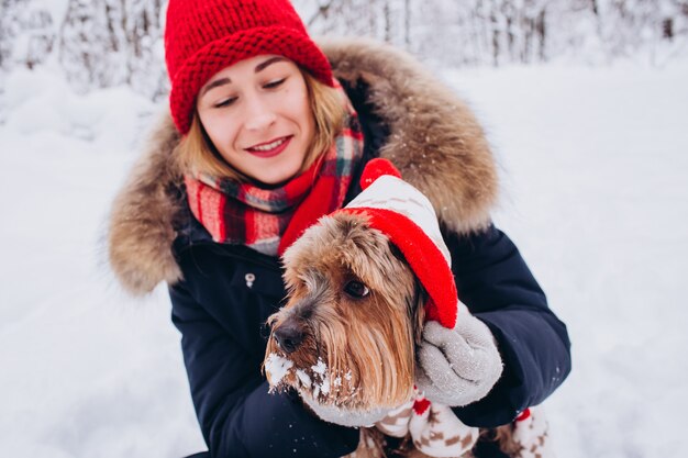 Closeup portrait of a girl with a dog in the winter forest, Dog in a red overalls, Girl in a red knitted hat
