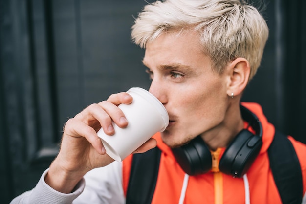 Closeup portrait of handsome Caucasian blond man with backpack and headphones relaxing during coffee break enjoy a cup of coffee outdoor against dark background in the city Lifestyle people concept