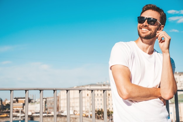 Closeup portrait of handsome smiling  hipster lambersexual modelStylish man dressed in white Tshirt Fashion male posing behind blue sky on the street background in sunglasses