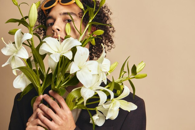 Photo closeup portrait of handsome tanned curly man in trendy sunglasses hold lilies near face posing isolated on over beige background fashion new collection offer retro style concept free place for ad