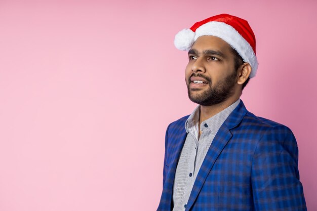 Closeup portrait of a handsome young indian man in a red Santa hat and a blue jacket smiling on isolated pink background with copy space.