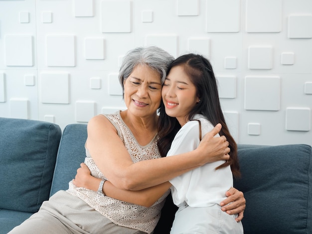 Closeup portrait of happy Asian senior mother or grandparent white hair embracing her beautiful daughter or grandchild with love care and comfort while sit on grey couch in living room at home