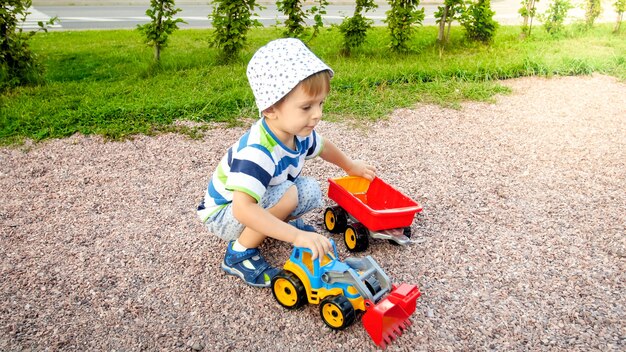 Closeup portrait of happy smiling 3 years old child boy digging sand on the playground with toy plastic truck or excavator