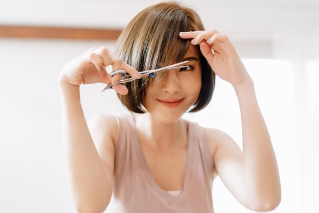 closeup portrait of happy young asian woman having her hair cut with scissors at home she's stay at home during the coronavirus pandemic self hair care during quarantine