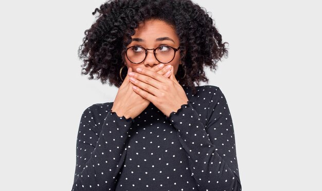 Photo closeup portrait of horrified african american young woman covering mouth with hands feeling scared looking aside with bulging eyes posing over white wall afro female student with shocked face