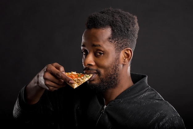 Foto ritratto del primo piano del ragazzo afroamericano affamato con la barba lunga con un pezzo di pizza gustosa in mano, mangiando fast food isolato su sfondo nero. cibo malsano, stile di vita, snack. vista laterale.