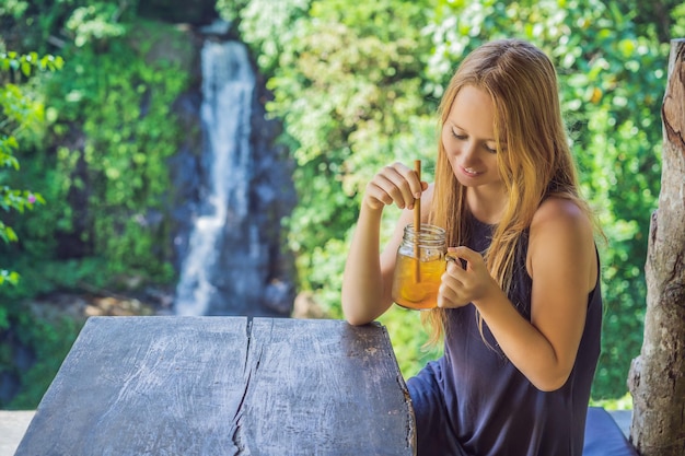 緑の自然と滝の庭の背景で幸せな気分でアイスティーを飲む美しい女性のクローズアップの肖像画の画像