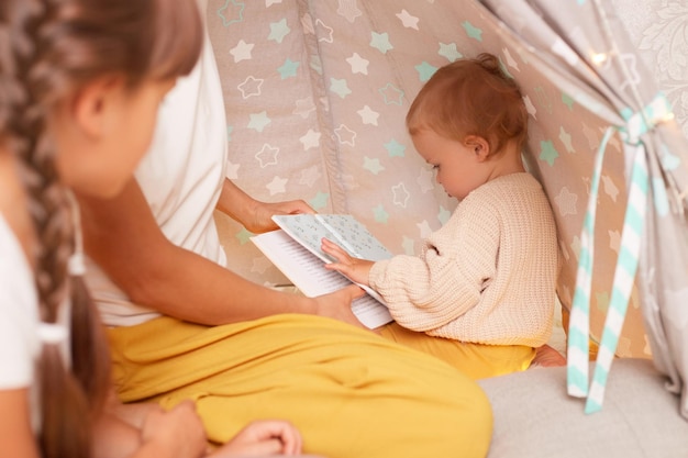 Photo closeup portrait of infant baby posing in wigwam with her sister with braids and faceless mother sitting near her charming kid holding book in hands playing with her family in teepee tent