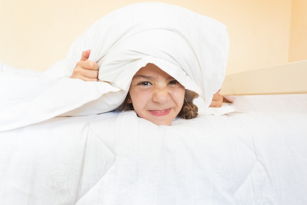 Closeup portrait of little angry girl lying in bed with pillow on head