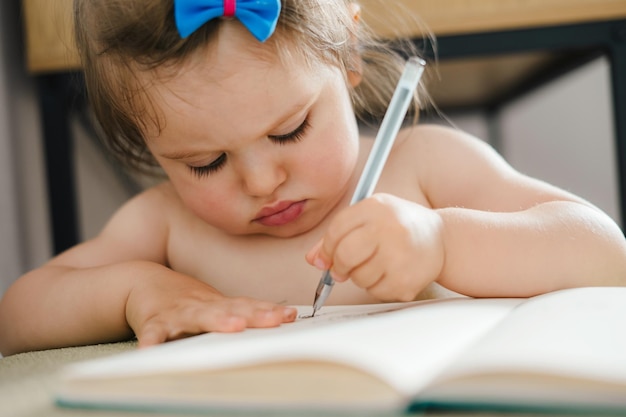 Closeup portrait of a little baby girl holding pencil drawing on notebook sitting at table at home Drawing kid activity Learning how to draw Creative arts and crafts