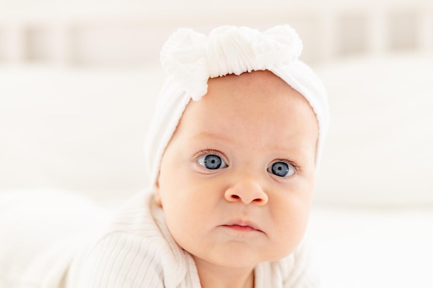 closeup portrait of a little baby girl with blue eyes in a white bodysuit on her tummy on a bed in a bright bedroom on a cotton bed a newborn baby girl healthy and happy smiling