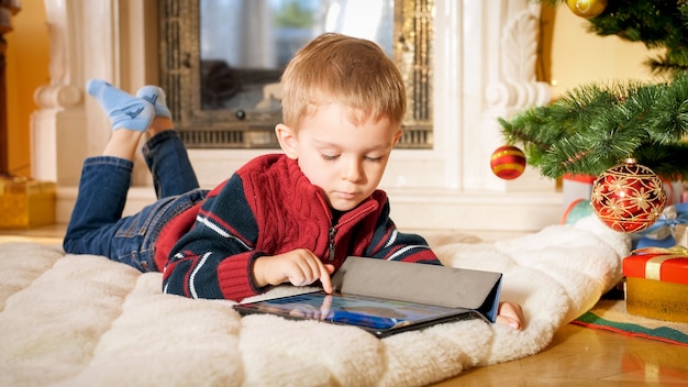 Closeup portrait of little boy lying on floor at living room decorated for Christmas and playing on digital tablet computer