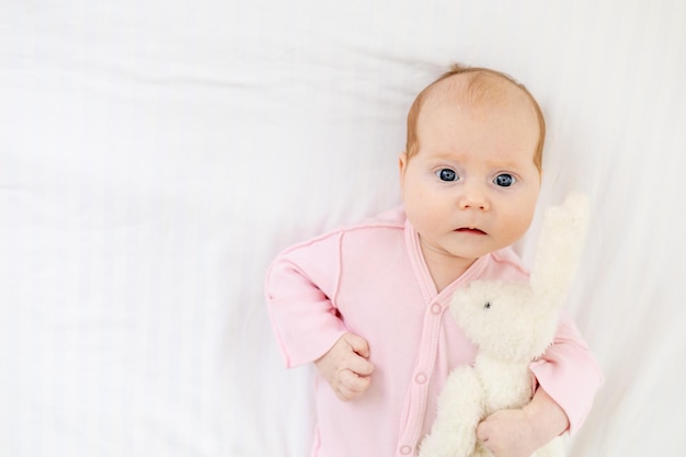 Closeup portrait little newborn baby girl with blue eyes lies on a white cotton bed at home and smiles hugging a plush hare