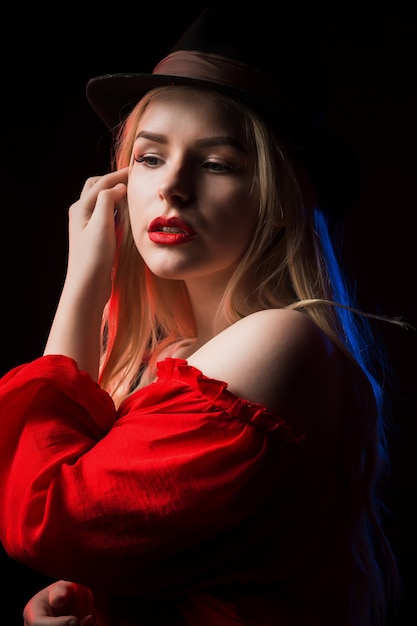 파란색과 빨간색 빛으로 그림자 속에서 포즈를 취하는 모자를 쓴 고급스러운 금발 여성의 클로즈업 초상화