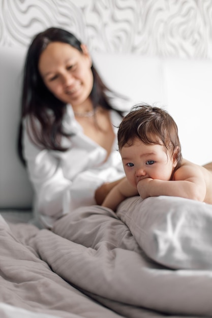 Closeup portrait of naked baby lying on tummy on gray bed free copy space Happy mother on blurred background watching her little daughter in bedroom Concept of maternal affection and childcare